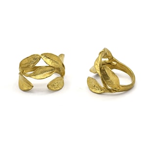 Puede incluir: Dos anillos dorados con forma de hojas. Los anillos tienen una superficie texturizada y están diseñados para parecer hojas. Los anillos se muestran sobre un fondo blanco.