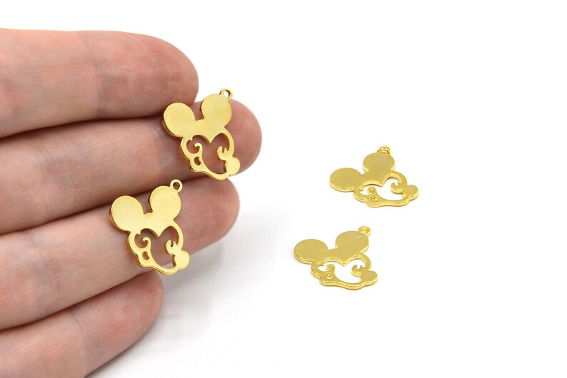 Puede incluir: Encantos de metal dorado con forma de cabeza de rat&oacute;n con remolinos decorativos. Los encantos tienen un peque&ntilde;o lazo en la parte superior para sujetarlos a joyas u otras manualidades. Varios encantos son visibles sobre un fondo blanco.