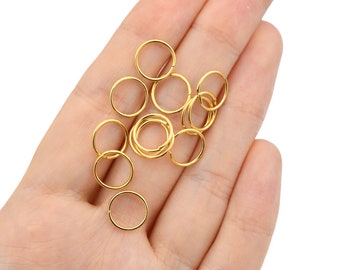 May include: A collection of small, gold-colored jump rings, likely for jewelry making, are held in a hand. The rings are circular and appear to be made of metal. The background is a plain white.