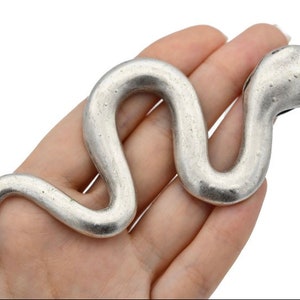Pode incluir: Uma escultura serpentina prateada. A cobra de metal é segurada em uma mão, seu corpo curvado em forma de S. A cabeça está ligeiramente levantada e a superfície tem um acabamento fosco texturizado. A escultura é provavelmente um item decorativo.