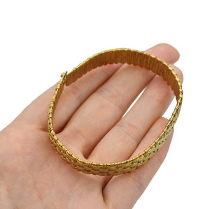 May include: A gold-colored bracelet with a woven design. The bracelet is held in a hand against a white background. The bracelet appears to be made of metal and has a clasp closure.