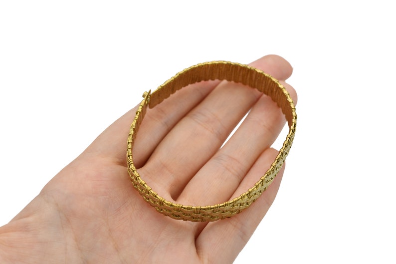 May include: A gold-colored bracelet with a woven design. The bracelet is held in a hand against a white background. The bracelet appears to be made of metal and has a clasp closure.