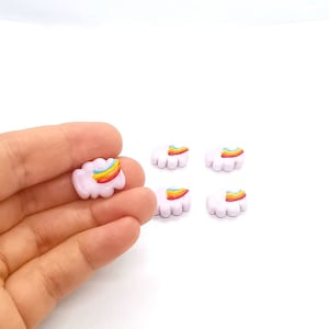 May include: Small, cloud-shaped charms with a rainbow design. The charms are light pink with a colourful rainbow arching across them. One charm is held in a hand, while four others are scattered on a white surface.
