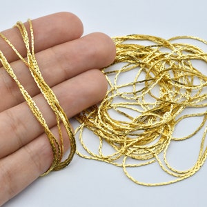 May include: A collection of gold-colored, flat, chain-like strands. The strands are intertwined and appear to be made of a metallic material. The chains are arranged in a loose, tangled pile, with some strands held in a hand.