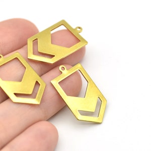 Könnte beinhalten: Drei goldfarbene geometrische Anhänger mit rechteckiger Form und einem Chevron-Design, das aus der Mitte herausgeschnitten ist. Jeder Anhänger hat oben eine kleine Schlaufe zum Befestigen an einer Kette oder einem anderen Schmuckbestandteil. Die Anhänger sind auf weißem Hintergrund.