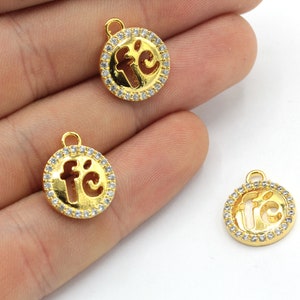 May include: Gold-colored round charm pendants with the word "fé" in a cut-out design. The pendants are encircled by a row of small, clear crystals. Each charm has a small loop at the top for attaching to a chain or bracelet.