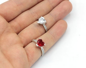 May include: Two heart-shaped rings. One ring features a clear, heart-shaped stone, while the other has a red heart-shaped stone. Both rings have a silver band with small, clear stones.