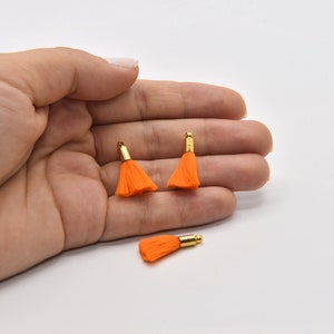 May include: Three small orange tassels with gold-colored metal caps. Two tassels are upright, and one is lying down. The tassels are on a white background. These could be used for jewelry making or other craft projects.