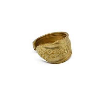 May include: A gold-colored ring with a floral pattern. The ring appears to be made from a spoon, with the spoon's bowl forming the decorative front. The ring is open at the back, allowing for size adjustment.