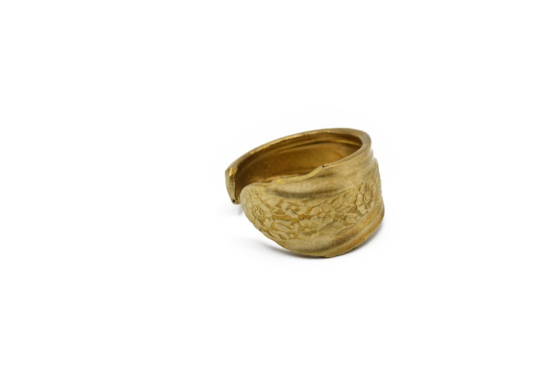 May include: A gold-colored ring with a floral pattern. The ring appears to be made from a spoon, with the spoon's bowl forming the decorative front. The ring is open at the back, allowing for size adjustment.