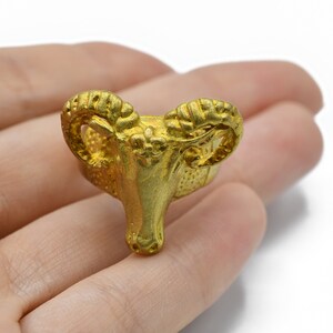 May include: A gold-coloured ram head figurine with detailed horns and facial features. The ram is a symbol of strength and leadership. The figurine is held in a hand against a white background.