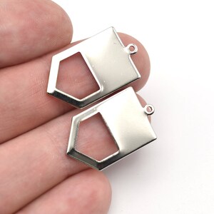 Może przedstawiać: Dwa srebrne metalowe zawieszki. Każda zawieszka ma geometryczny kształt z prostokątną górą i pięciokątnym dołem z wycięciem. Mała pętla u góry do mocowania biżuterii.