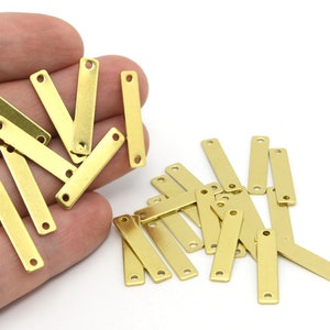 May include: A collection of small, rectangular brass metal tags with two holes at each end. The tags are a warm gold color and are scattered on a white background. These tags are ideal for jewelry making or crafting.