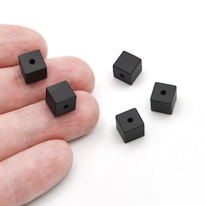 May include: Five small, black square beads with a hole drilled through the centre of each. The beads are scattered on a white surface. The beads are approximately 0.8 cm in size.