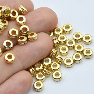 May include: A collection of small, gold-colored metal beads. Each bead is cylindrical with a hole running through the center. The beads have a shiny, metallic finish and are scattered across a light background.