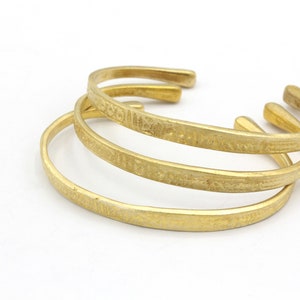 May include: Three gold-colored cuff bracelets with a textured surface are displayed against a white background. The bracelets are open-ended and appear to be made of metal. The bracelets are stacked on top of each other.