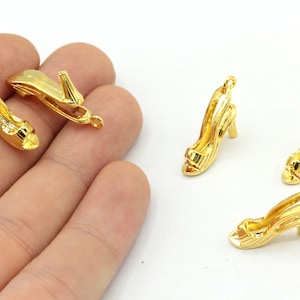 May include: A collection of small, gold-coloured high heel shoe charms. The charms feature a detailed design, including a strap across the toes and a small heel. Each charm has a loop at the top for attaching to jewellery or other crafts.