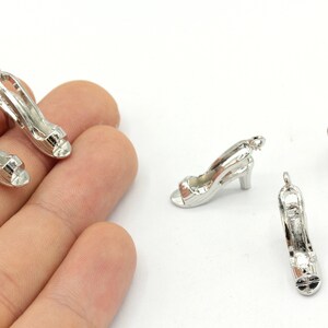 May include: Silver high heel shoe charms. These small, detailed charms feature an open-toe design with a strap and a small heel. The charms are likely made of metal and are suitable for jewellery making or crafting.