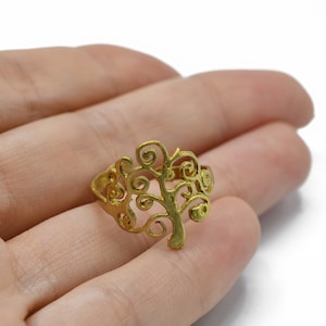 Op de afbeelding: Een goudkleurige ring met een boomontwerp. De boom van de ring heeft een stam en takken met wervelende details. De ring wordt in een hand gehouden tegen een witte achtergrond. De ring is een sieraad.