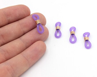May include: Four lavender-colored eyeglass retainers with gold-colored wire accents. The retainers are made of a flexible material and are designed to attach to the arms of eyeglasses to keep them secure. The retainers are shown against a white background.
