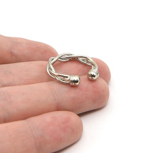 May include: A silver-colored ring with a braided design. The ring has a unique open design with two rounded ends. The ring is held in a hand against a white background. The ring is a piece of jewelry.
