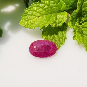Op de afbeelding: Een ovaalvormige, gefacetteerde robijn met een diepe, verzadigde magenta-rode kleur. De steen heeft een gepolijst oppervlak met zichtbare interne insluitingen en geometrische facetten die het licht reflecteren, geplaatst bij levendige groene muntblaadjes.