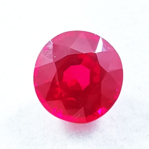 May include: A round, faceted gemstone with a vibrant, saturated red color. The stone features a brilliant cut with multiple triangular and kite-shaped facets that reflect light, creating a clear, polished surface with sharp edges.