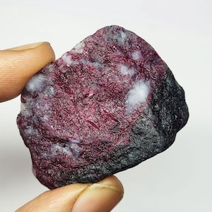 Peut inclure: Une pierre précieuse brute, d'un rouge bordeaux profond, parsemée de taches blanches et bordée d'une couleur sombre, presque noire. La surface de la pierre présente un aspect texturé et irrégulier, révélant des formations minérales naturelles.