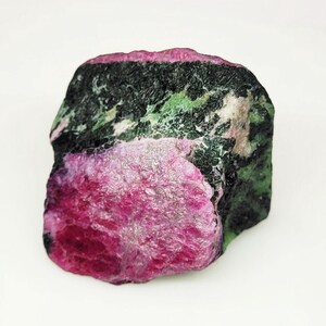 Può includere: Una gemma grezza con un vivace mix di colori. La pietra presenta una tonalità magenta intenso su un lato, che sfuma in un motivo maculato verde scuro e nero. La forma generale è irregolare e naturale.