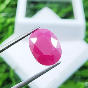 May include: A vibrant, oval-shaped, faceted pink gemstone held by metal tweezers. The stone features a polished surface with multiple geometric facets that reflect light, showcasing a deep, saturated magenta-pink color throughout the gem.
