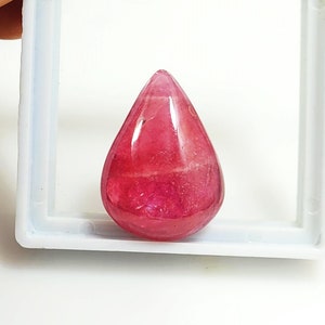 May include: A teardrop-shaped, polished gemstone in shades of pink and red. The gem is set in a white plastic holder, and the stone's surface reflects light, showcasing its translucent quality. The gem is likely for jewelry making.