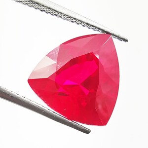 May include: A vibrant, trillion-cut red gemstone held by metal tweezers. The stone features a triangular shape with multiple facets that reflect light, creating a brilliant, saturated crimson appearance against a plain white background.