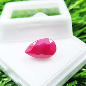 Puede incluir: Una gema de rubí facetada en forma de pera, exhibida en una caja de plástico blanco. El rubí es de un color rojo intenso y vibrante, con una superficie pulida y reflectante. La caja se encuentra sobre un fondo de hierba verde.
