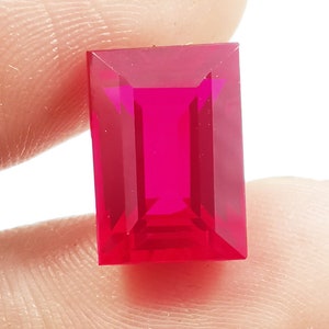 Könnte beinhalten: Ein leuchtender, rechteckig geschliffener Edelstein in einem tiefen, gesättigten Magentarot. Der Stein besitzt eine facettierte Oberfläche, die das Licht reflektiert und scharfe geometrische Muster sowie eine klare Tiefe in der transparenten Struktur erzeugt.