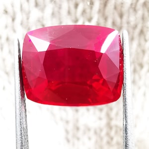 May include: A vibrant, cushion-cut red gemstone held by silver-toned metal tweezers. The stone features a faceted surface that reflects light, showcasing a deep, saturated crimson hue throughout its transparent, polished crystalline structure.