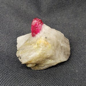May include: A rough, uncut gemstone featuring a vibrant red crystal atop a light-colored, textured rock. The crystal is the focal point, showcasing its natural, unpolished form. The stone sits against a dark gray background.
