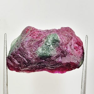 Puede incluir: Una piedra preciosa en bruto y sin pulir con una paleta de colores magenta intenso y rojo carmesí, que presenta una prominente inclusión de color verde oscuro texturizado en el centro. La piedra tiene una forma irregular.