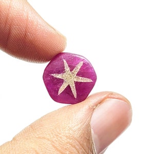以下が含まれることがあります： 白星模様の、六角形にカットされたマゼンタ色の宝石。白い背景を背景に、2本の指で宝石を持っています。星のデザインは8つのポイントがあります。