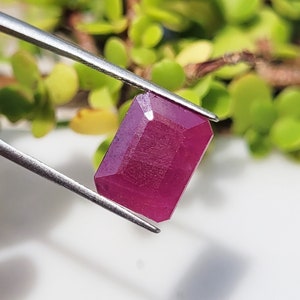 May include: A rectangular faceted gemstone with a deep magenta-red colour held by silver-toned metal tweezers. The stone features a clear, polished surface with sharp edges and geometric facets that reflect light across its vibrant, saturated surface.