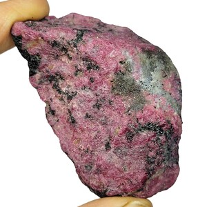 Könnte beinhalten: Ein roher Rubin-Edelstein mit einem tiefrosa Farbton, gesprenkelt mit schwarzen und grauen Einschlüssen. Das rohe Mineral wird zwischen Fingern gehalten, wodurch seine unregelmäßige Form und strukturierte Oberfläche sichtbar werden. Der Edelstein ist vor einem weißen Hintergrund platziert.