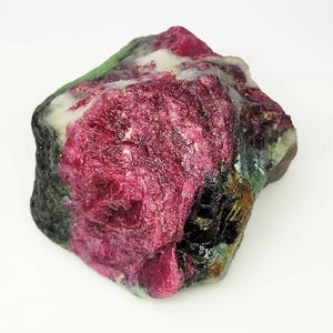 May include: A raw ruby gemstone, displaying a rich red colour with patches of black, green, and white. The natural stone has a textured surface and an irregular shape, highlighting its natural beauty.
