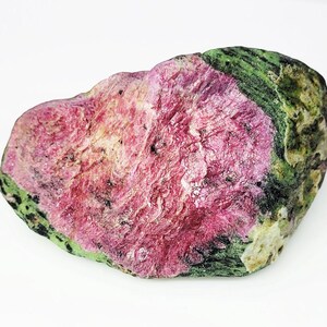Op de afbeelding: Een ruw geslepen edelsteen met een levendig roze centrum en een groene buitenrand. De steen heeft een textuur met zichtbare minerale formaties. De kleuren zijn natuurlijk en aards, wat wijst op een ruwe, ongepolijste staat.