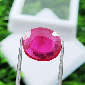 Puede incluir: Una gema de rubí facetada, de forma ovalada, sujeta con pinzas plateadas. El rubí es de un color rojo intenso y vibrante, con múltiples facetas que reflejan la luz. El fondo es verde y blanco suaves.