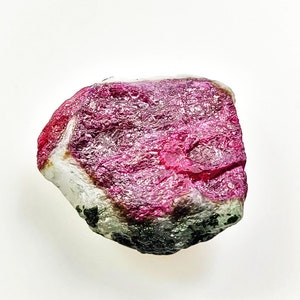 以下が含まれることがあります： 鮮やかなマゼンタピンクの結晶中心部を持つ、未研磨の原石。表面は粗く質感があり、端に向かって淡い半透明の白へと変化し、底部は黒とグレーの斑点模様の鉱物インクルージョンが見られる不規則な形状の天然石です。