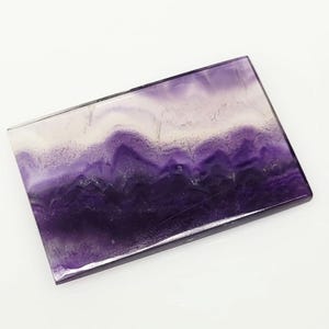 Puede incluir: Cristal rectangular de fluorita con apariencia en capas. La piedra presenta bandas de tonos blanco, morado claro y morado oscuro. La superficie pulida refleja la luz, mostrando los patrones y colores naturales del mineral.