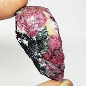 Könnte beinhalten: Ein rohes, unpoliertes Mineralexemplar mit einer Kombination aus tief magentaroten kristallinen Abschnitten, dunkelgrünen faserigen Einschlüssen und Flecken aus opakem weißem Material, das eine gezackte, unregelmäßige Oberfläche aufweist.
