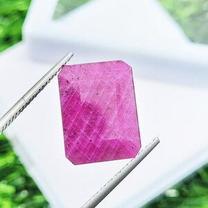 Puede incluir: Una piedra preciosa rectangular facetada con un tono rosa magenta vibrante, que presenta una superficie pulida con inclusiones internas visibles y motas oscuras por toda la piedra, sostenida por unas pinzas de metal plateado.