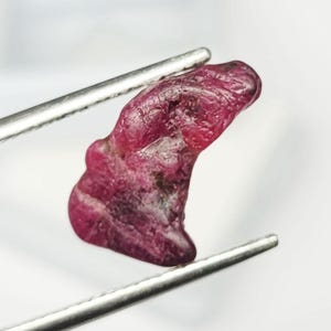 May include: A raw, unpolished ruby gemstone with a deep, saturated magenta-red hue and an irregular, organic shape. The stone features a textured, pitted surface with natural variations in color intensity and is held by metal tweezers.