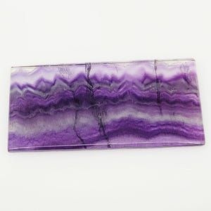 Puede incluir: Una pieza rectangular de fluorita con un patrón en capas y ondulado. La piedra presenta tonos de morado, lavanda y blanco, con vetas de color gris oscuro. La superficie pulida refleja la luz, resaltando las variaciones naturales del mineral.