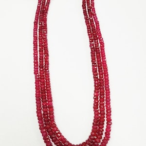 May include: A multi-strand necklace featuring three rows of faceted, deep red gemstone beads. The strands are gathered at the top with gold-colored metallic thread accents and red cord ties, creating a layered, uniform appearance.